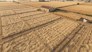 Vue aérienne réaliste d’une exploitation agricole française touchée par la sécheresse, avec des parcelles couvertes de paillage, des lignes de goutte-à-goutte et une petite citerne de récupération d’eau de pluie.